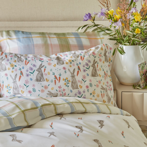 Bed with Easter bedding with bunnies, carrots and flowers on it. On the bedside table, there is a vase with flowers in it.