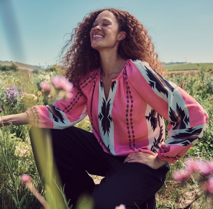 Woman in a field wearing a pink aztec print shirt with long sleeves and jeans.