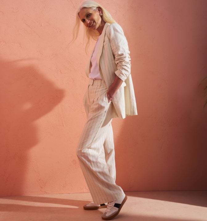 Woman wearing a modest workwear outfit consisting of a cream pinstripe blazer and wide leg trousers suit with a white t shirt and white trainers.