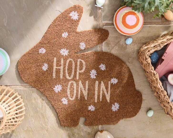 Easter doormat that is bunny shaped with the words 'hop on in' on it.